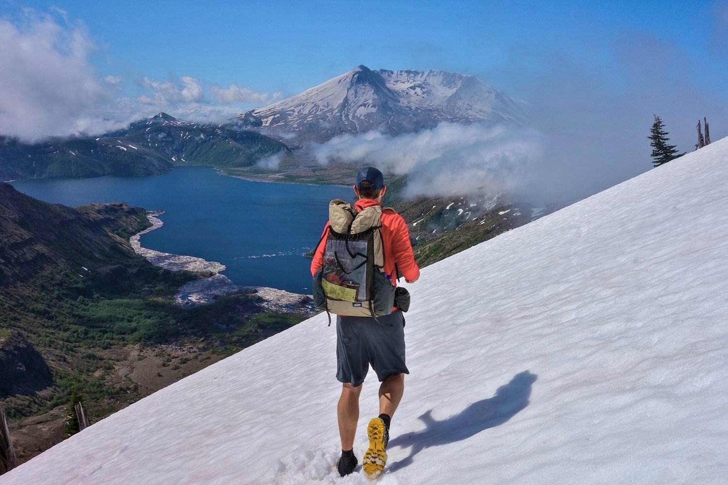 A hiker walking through a snow field in Nike Dri FIT Challenger Shorts with a mountain and lake view in the background
